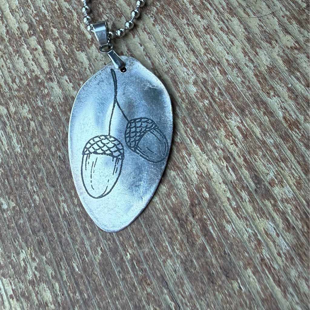 A silver-toned metal pendant necklace featuring a stamped acorn design, resting on a rustic wooden surface.