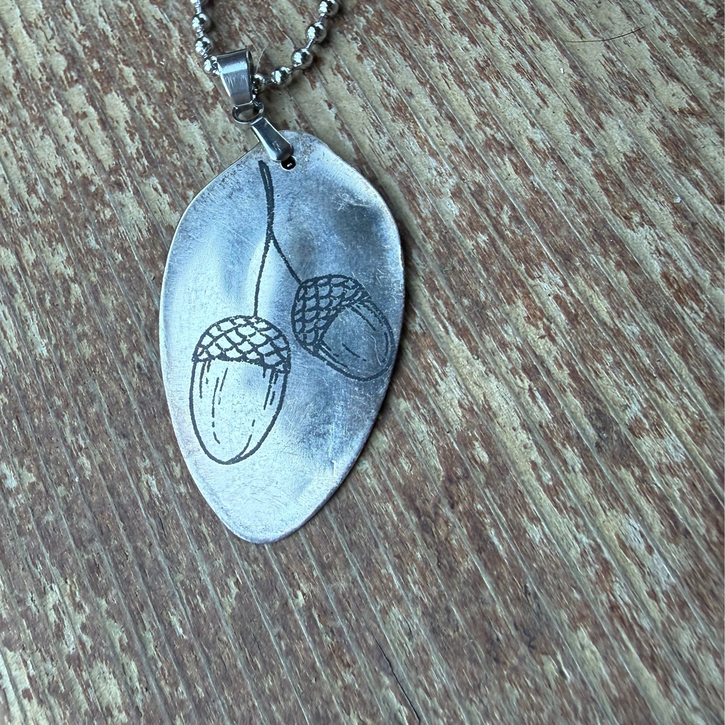 A silver-toned metal pendant necklace featuring a stamped acorn design, resting on a rustic wooden surface.