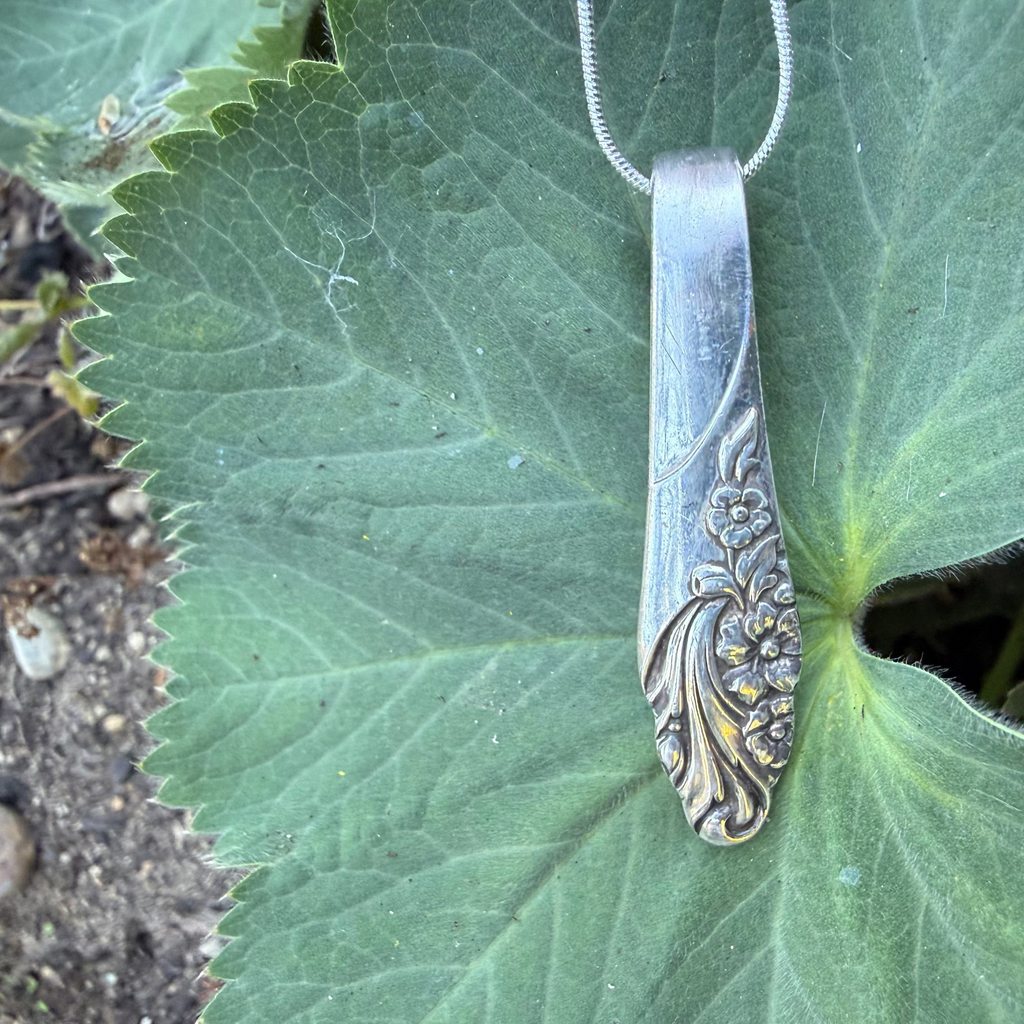 A vintage silver-toned metal pendant or charm with an ornate floral engraved handle, resting on a large green leaf outdoors.