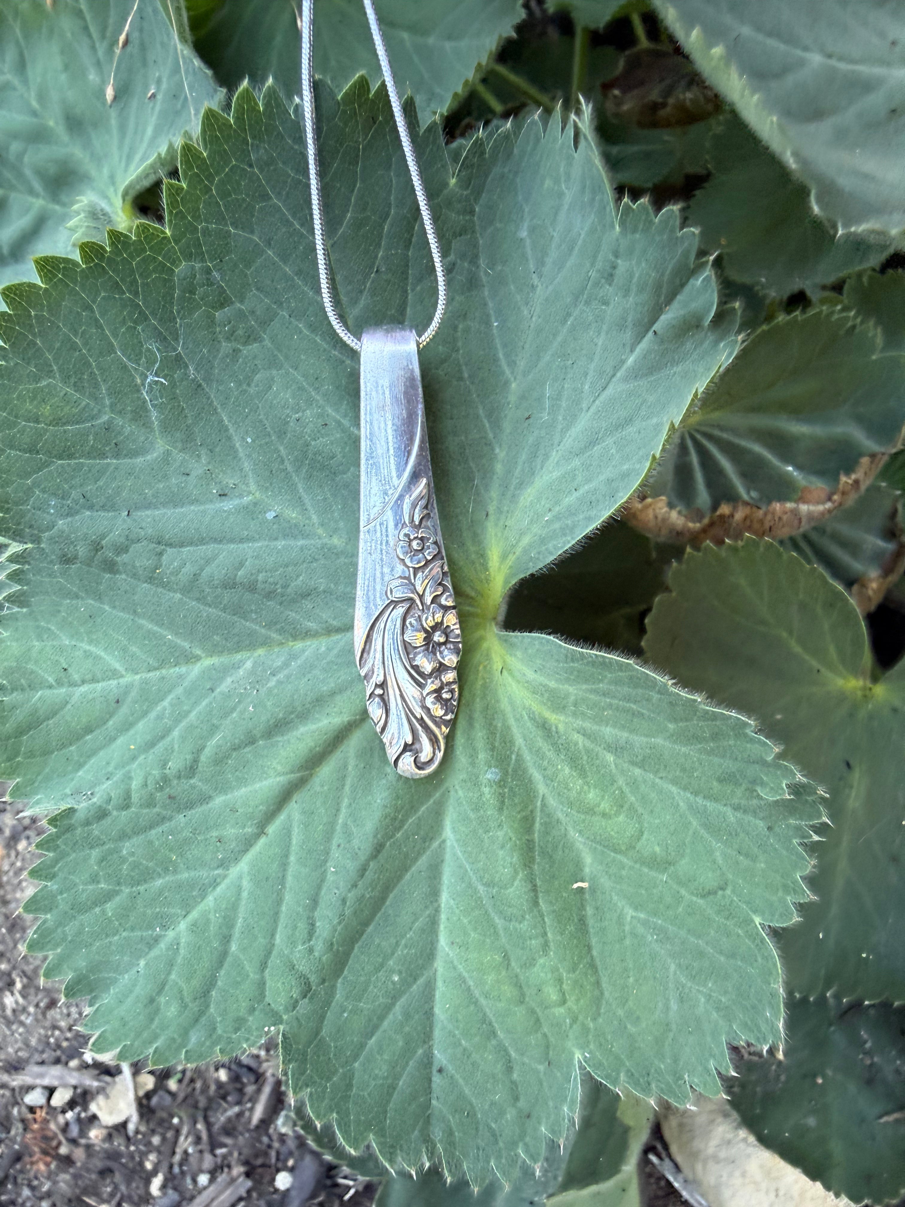 A vintage silver spoon pendant necklace with floral engravings, hanging from a silver chain against a green leaf background.