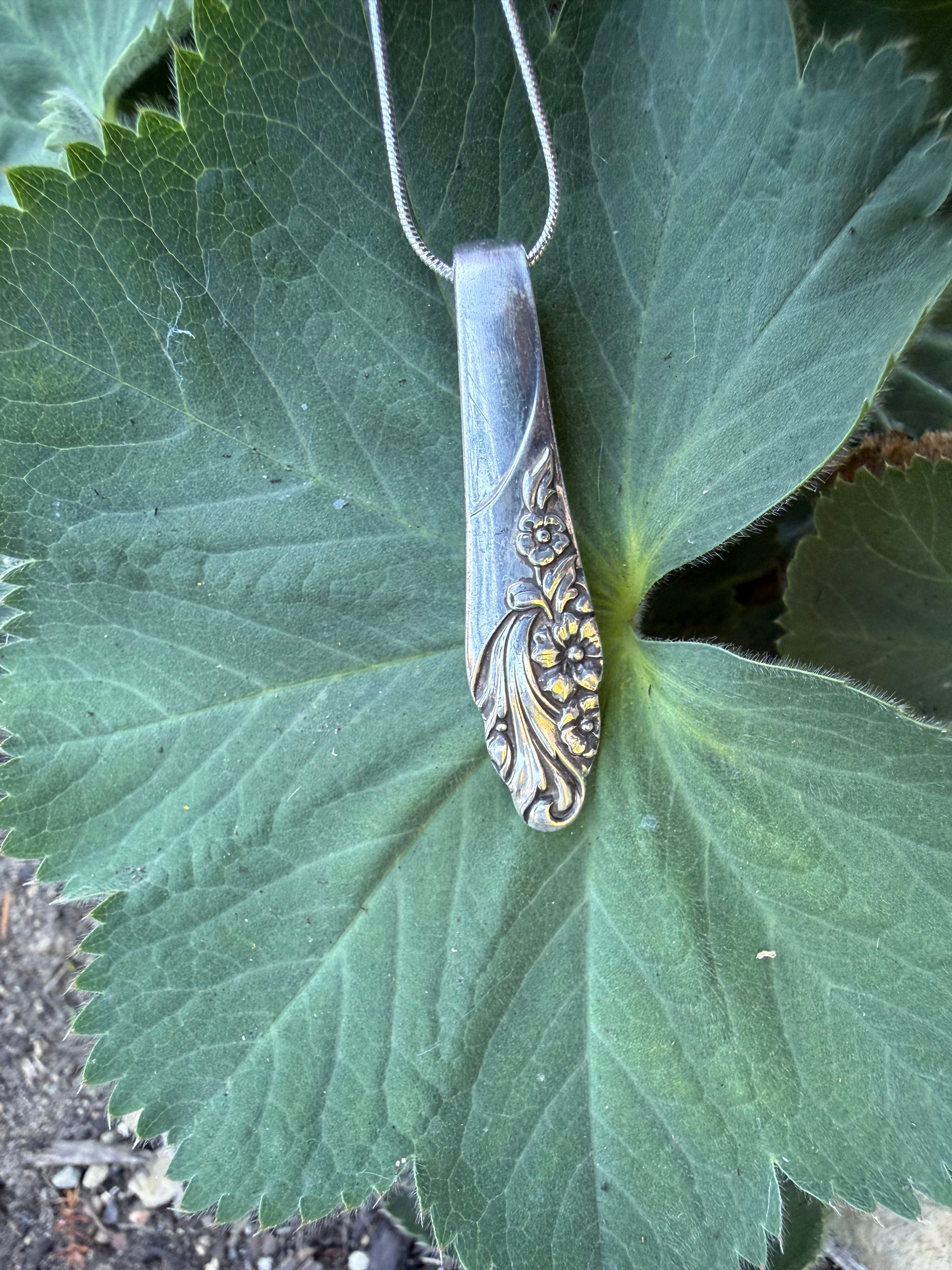 A vintage silver spoon pendant necklace with gold floral detailing, hanging from a silver chain against a large green leaf.