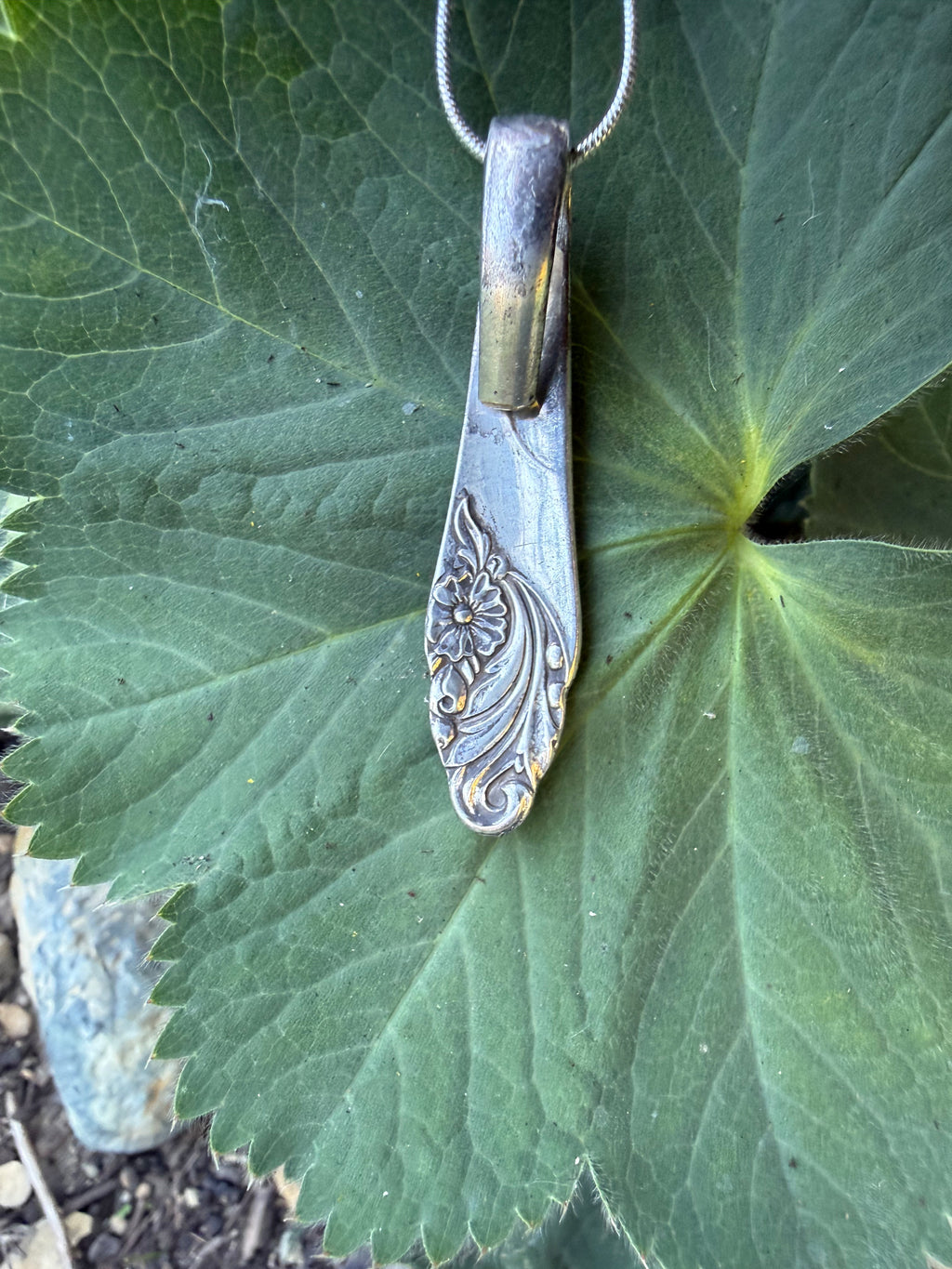 A vintage-style sterling silver spoon pendant with floral engravings, hanging from a silver chain against a large green leaf.