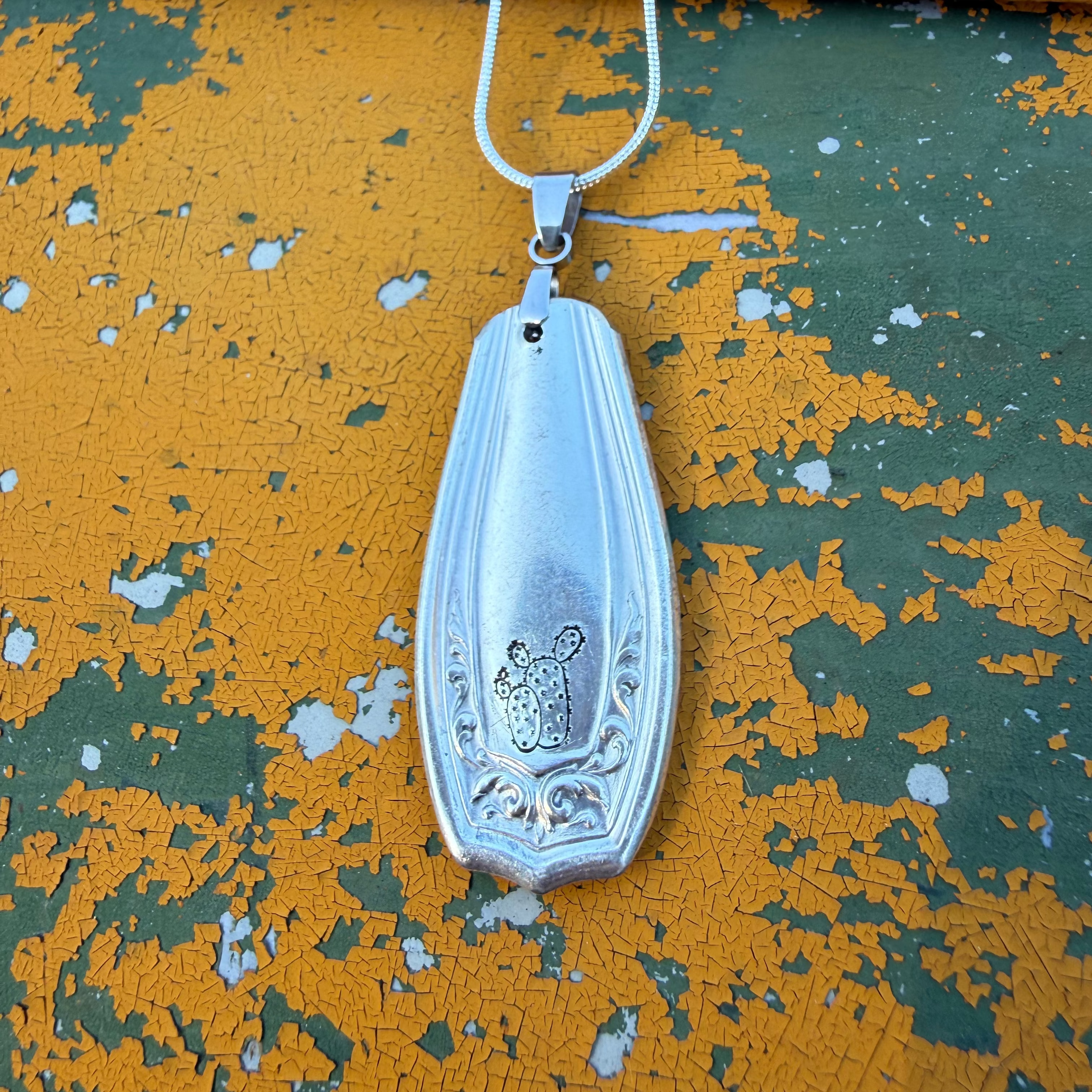A silver-toned metal pendant necklace with a vintage-style elongated pendant and a small central animal motif, hanging against a distressed yellow and green painted surface.