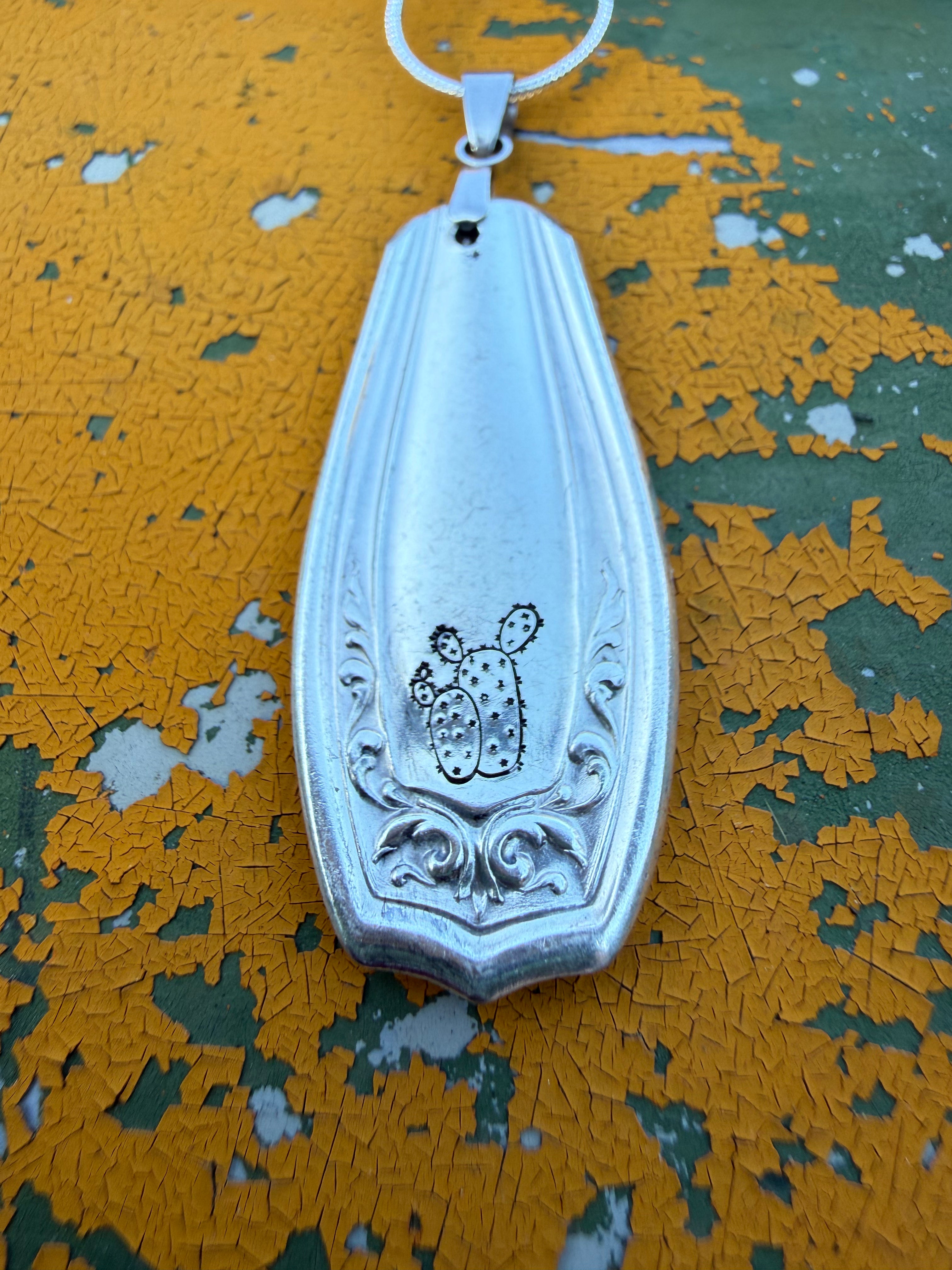 A silver-toned metal pendant with a cactus and scroll engraving, hanging from a matching silver chain against a distressed yellow and green surface.