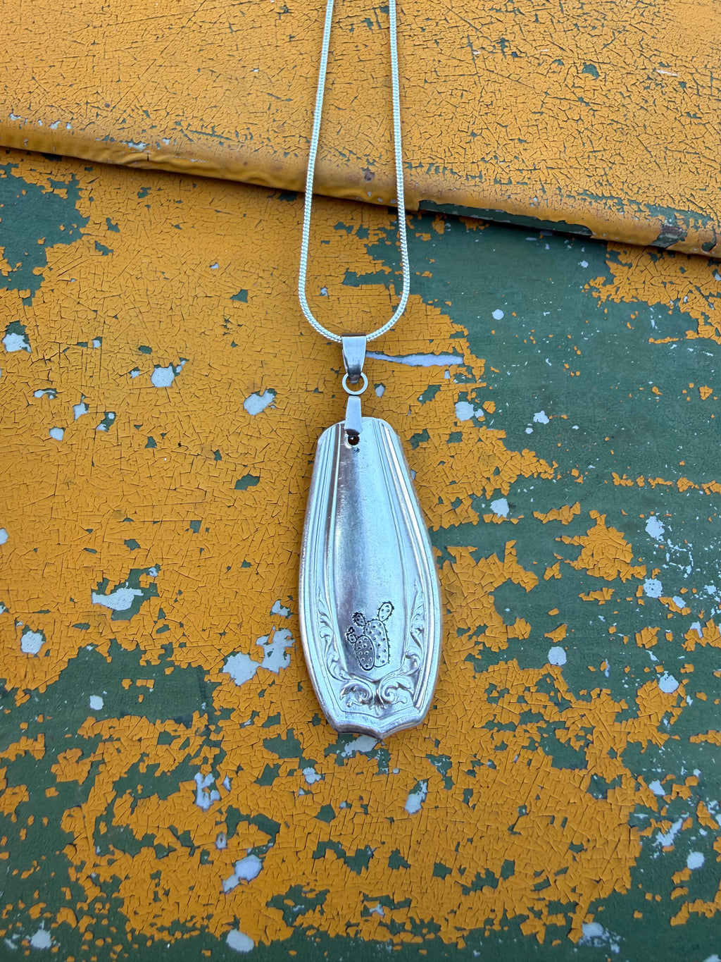 A silver pendant necklace with a vintage-style spoon ornament featuring a floral engraving, hanging against a distressed yellow and green painted metal background.