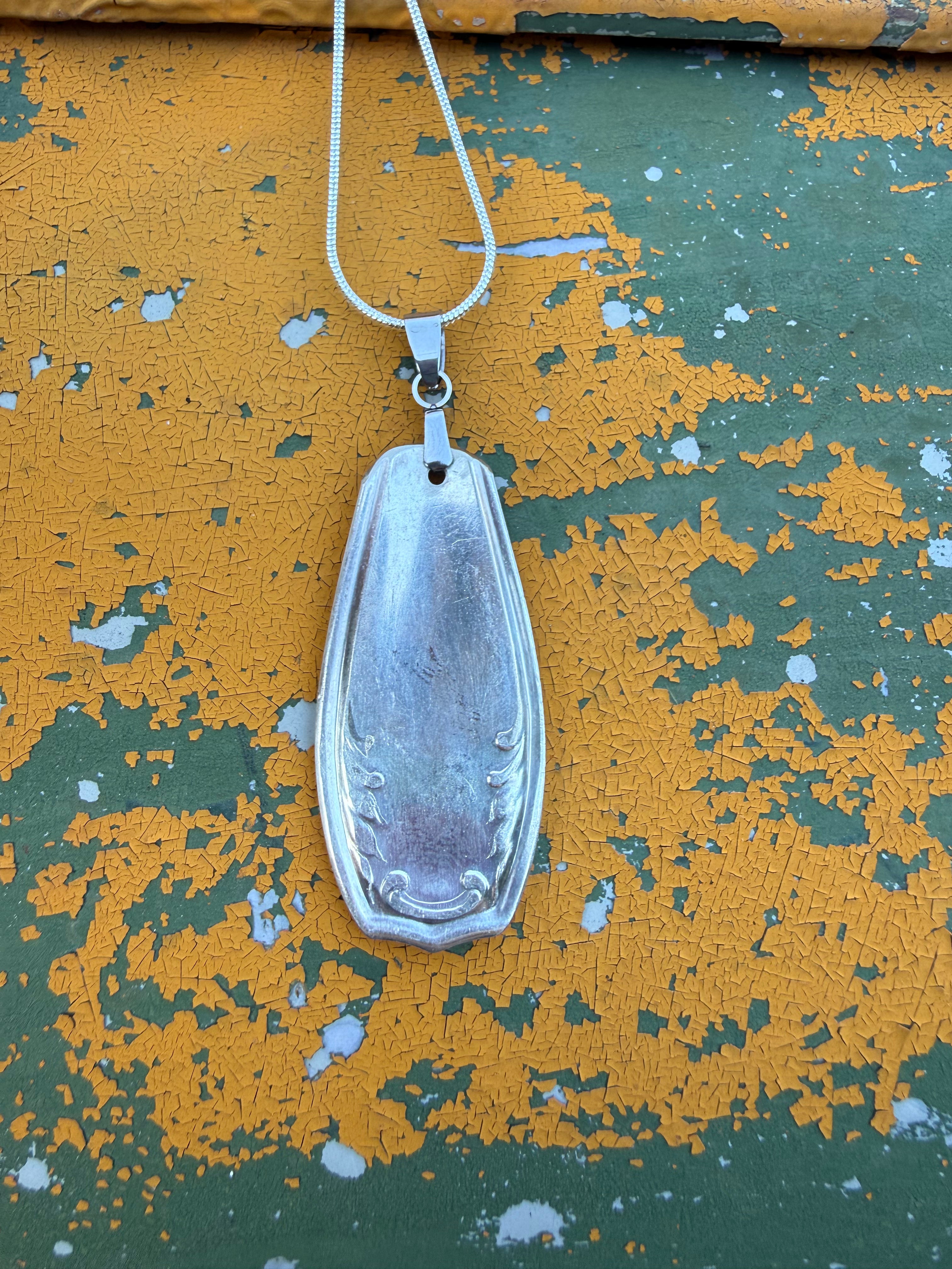 A silver pendant necklace with a teardrop-shaped engraved pendant on a silver chain, displayed against a distressed yellow and green painted wooden background.