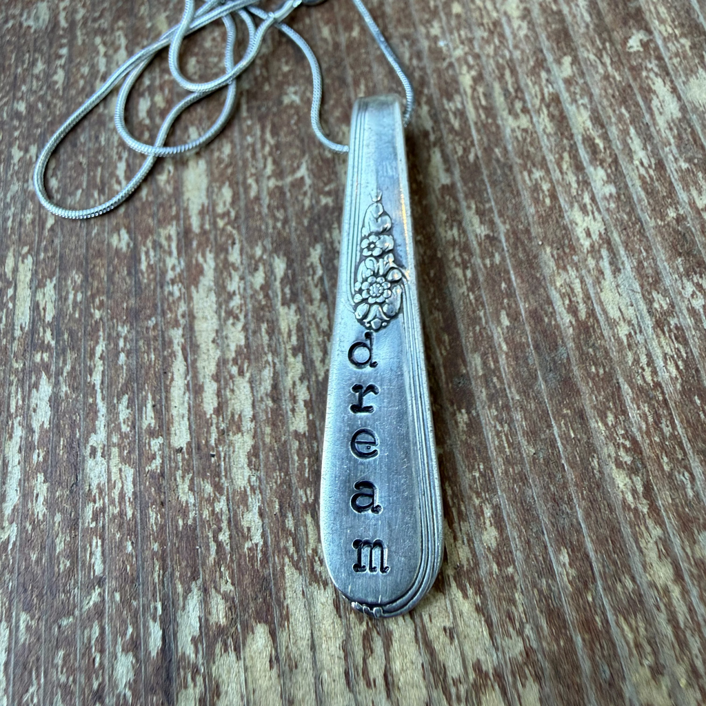 A silver spoon pendant necklace with the word 'dream' hand-stamped vertically, resting on a rustic wooden surface.