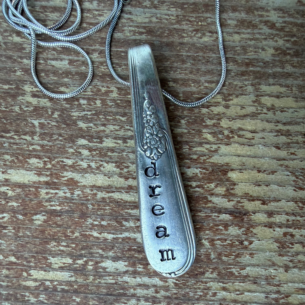 A vintage sterling silver spoon pendant engraved with the word 'dream' and a floral motif, displayed on a weathered brown wooden surface with its matching snake chain.