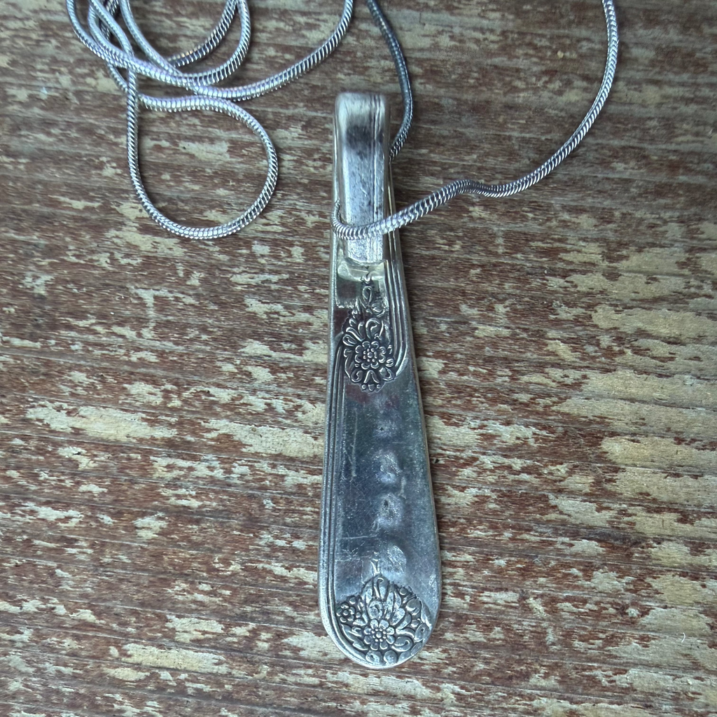 A vintage sterling silver spoon pendant necklace with floral engravings on the handle, resting on a weathered, peeling brown wood surface.