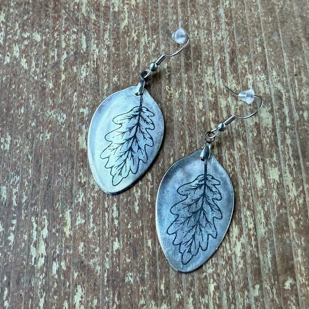 A pair of handmade silver leaf dangle earrings with black leaf engravings, displayed on a rustic brown wooden surface.