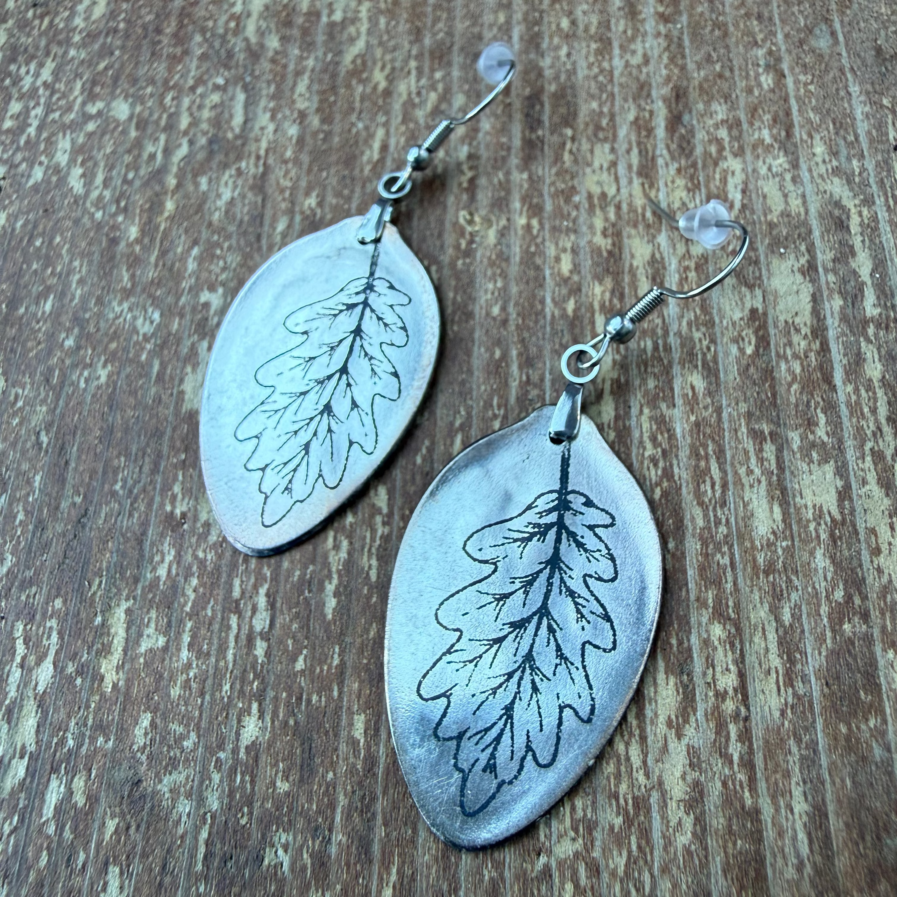 A pair of silver-toned dangle earrings shaped like oak leaves with engraved leaf vein details, shown on a rustic wooden surface.