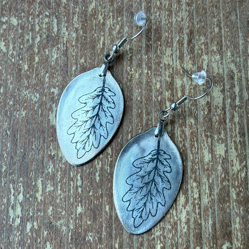 A pair of handmade dangle earrings with oval silver-toned metal pendants stamped with an oak leaf design, hanging from silver fishhook wires with small clear beads, displayed on a rustic wooden surface.