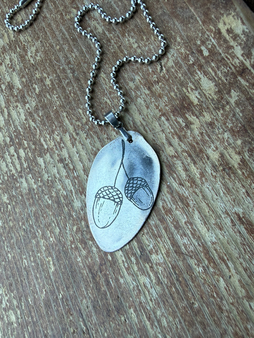 A silver-toned metal pendant necklace featuring an engraved acorn and acorn cup design, resting on a rustic wooden surface.